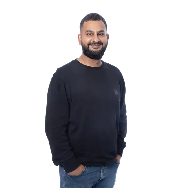 Smiling man in a black sweater and blue jeans, standing against a blurred light blue background.