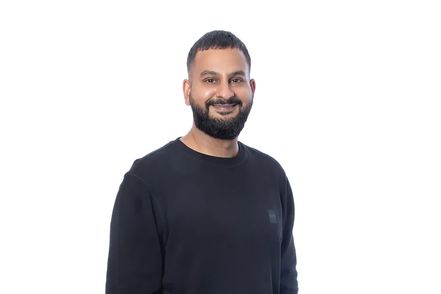 Smiling man in a black sweater and blue jeans, standing against a blurred light blue background.