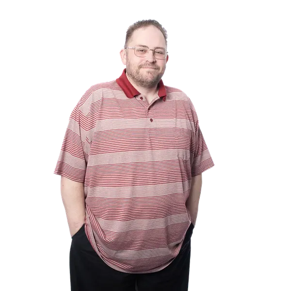 Man in a striped red polo shirt and black pants, standing with hands in pockets against a blurred background.