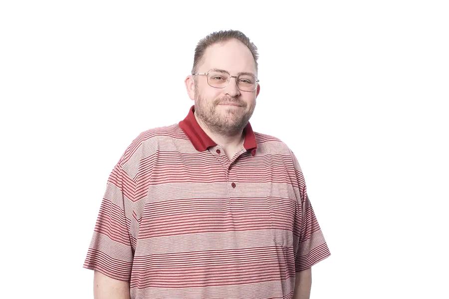 Man in a striped red polo shirt and black pants, standing with hands in pockets against a blurred background.