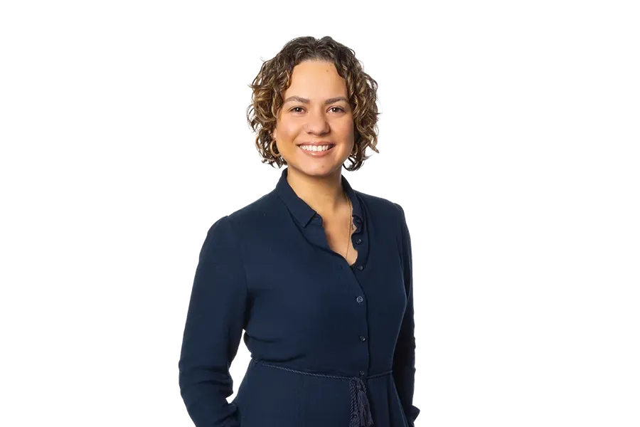Smiling person in a dark blue buttoned dress, hands in pockets, against a plain white background.