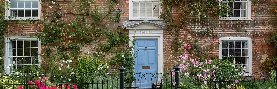 Charming brick house with a blue door, surrounded by climbing plants and a variety of colorful flowers behind a black wrought-iron fence.