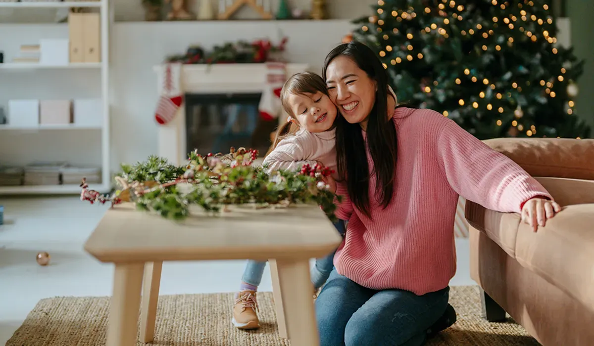Mother and daughter hug by a coffee table with a holiday wreath, a decorated Christmas tree and stockings in the background.