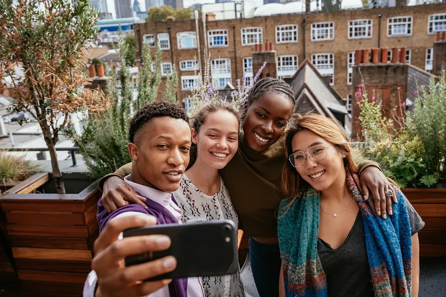 Four friends taking a selfie on a rooftop, smiling with arms around each other, buildings and greenery in the background.