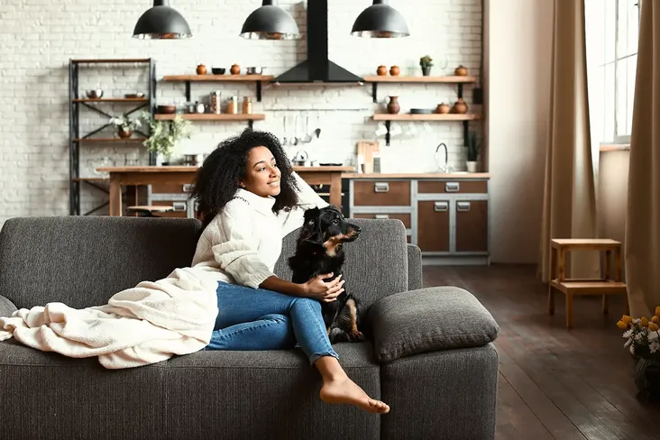 young woman sitting on the sofa with her small black dog, in her flat, covered with a blanket and smiling whilst looking out of the window. Behind is an open plan kitchen with a white brick wall and modern shelves. She's excited as she is leaving her rented flat and moving into her first home