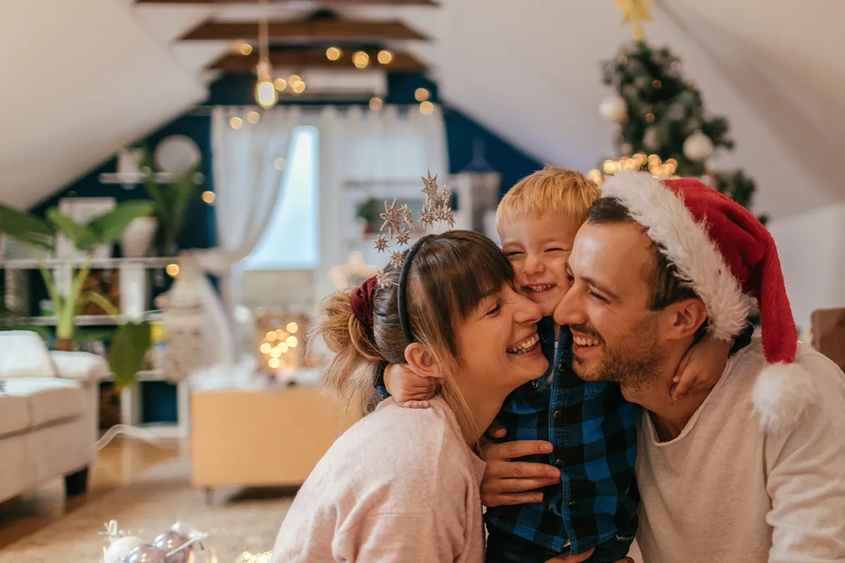 Parents and young son laughing and hugging in a cozy living room with a decorated Christmas tree; father wears a Santa hat.