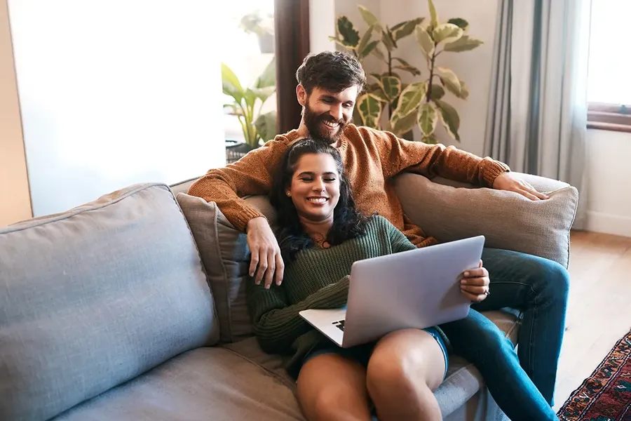 young couple of first time buyers sitting on a sofa in a living room, looking at a laptop and smiling. Researching if they might not have to pay stamp duty land tax when buying a house