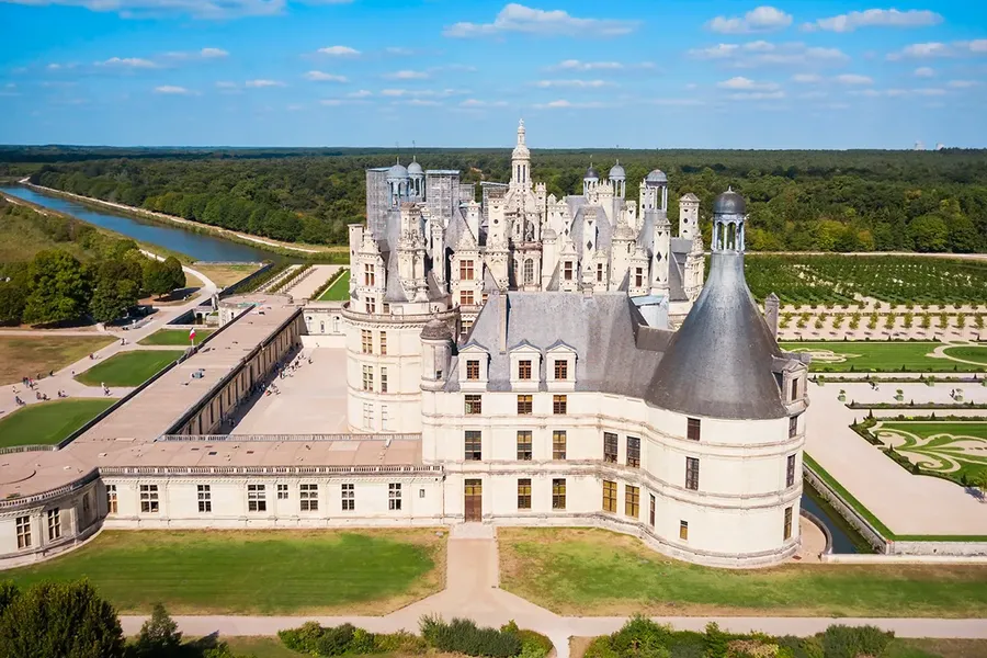 aerial view of a stunning french chateau with silver roof and turrets and white painted bricks. There's a forest behind it and a long lake.