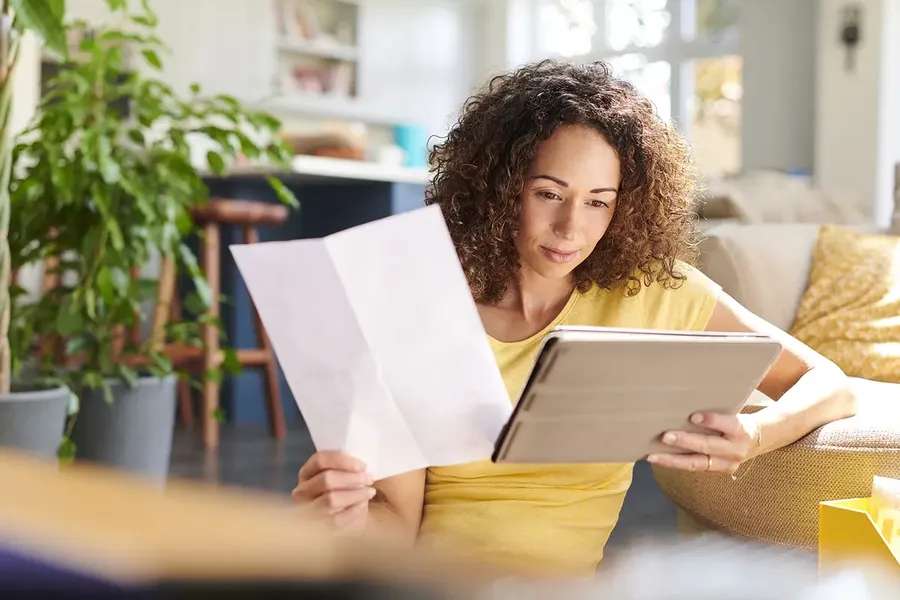 Woman with curly hair in a yellow shirt reading documents and a tablet while sitting on a couch in a bright living room.