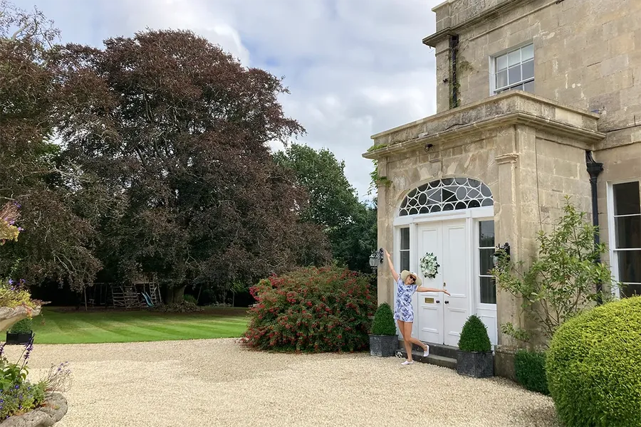 the winner of the my home move mansion tester competition dancing outside the large white front door of a yellow stoned old mansion with gravel drive and perfect gardens
