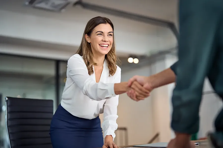 Smiling businesswoman in white blouse and navy skirt leaning over a desk to shake hands with a colleague in a modern office.