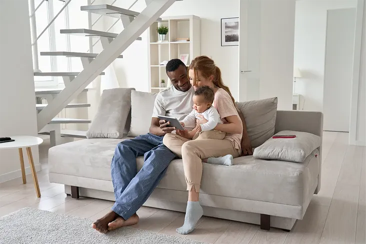 happy family of a man, woman and young baby sat together on a sofa in their rented apartment looking at house moving sites on their tablet to find their perfect home
