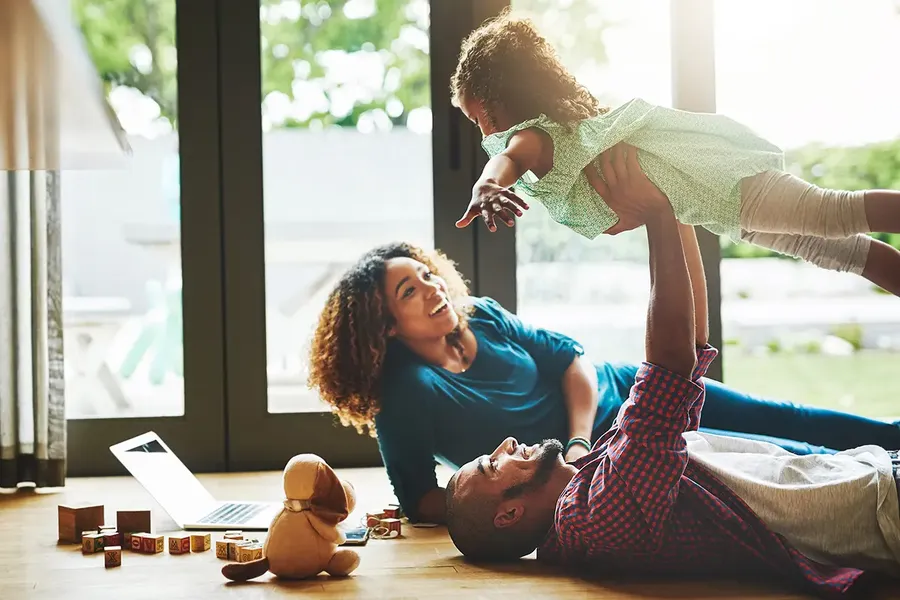 family of 3 lying on the floor of their new home. the dad is lifting the daughter up into the air and everyone is smiling
