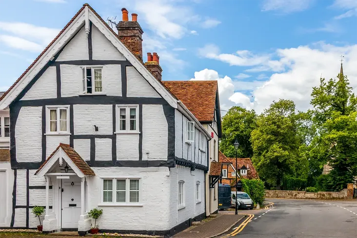 A white and black timber-framed grade-2 listed house on a quiet street, surrounded by greenery, with a church spire visible in the background.