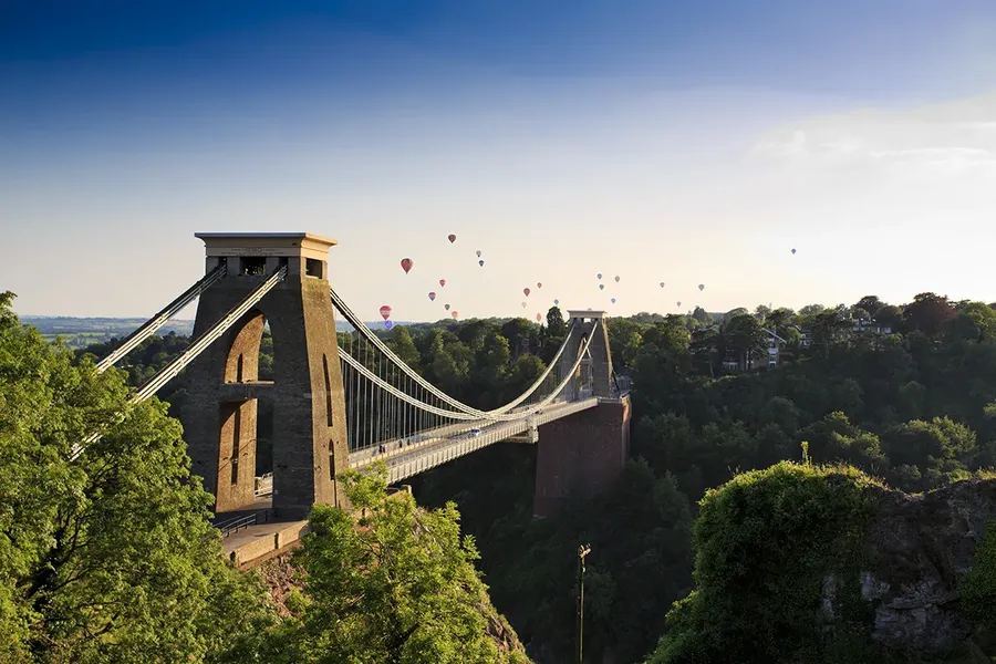 ariel view of the clifton suspension bridge in bristol. it is a clear day and the sky is filled with hot air balloons, the bridge is surrounded by leafy treetops