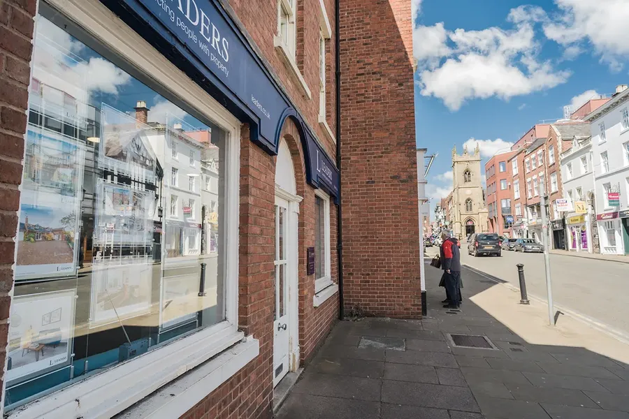 High street view with a real estate agency window displaying listings. A church is visible in the background under a blue sky with clouds.