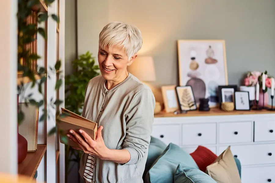 a mature homeowner standing in the window of her flat, looking at a guide to conveyancing getting ready to sell her house