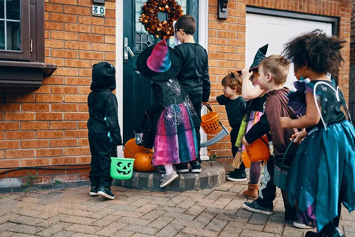 seven young children dressed up in halloween costumes, carrying treat buckets. The child at the front is knocking on the door of a redbrick house waiting for someone to answer.