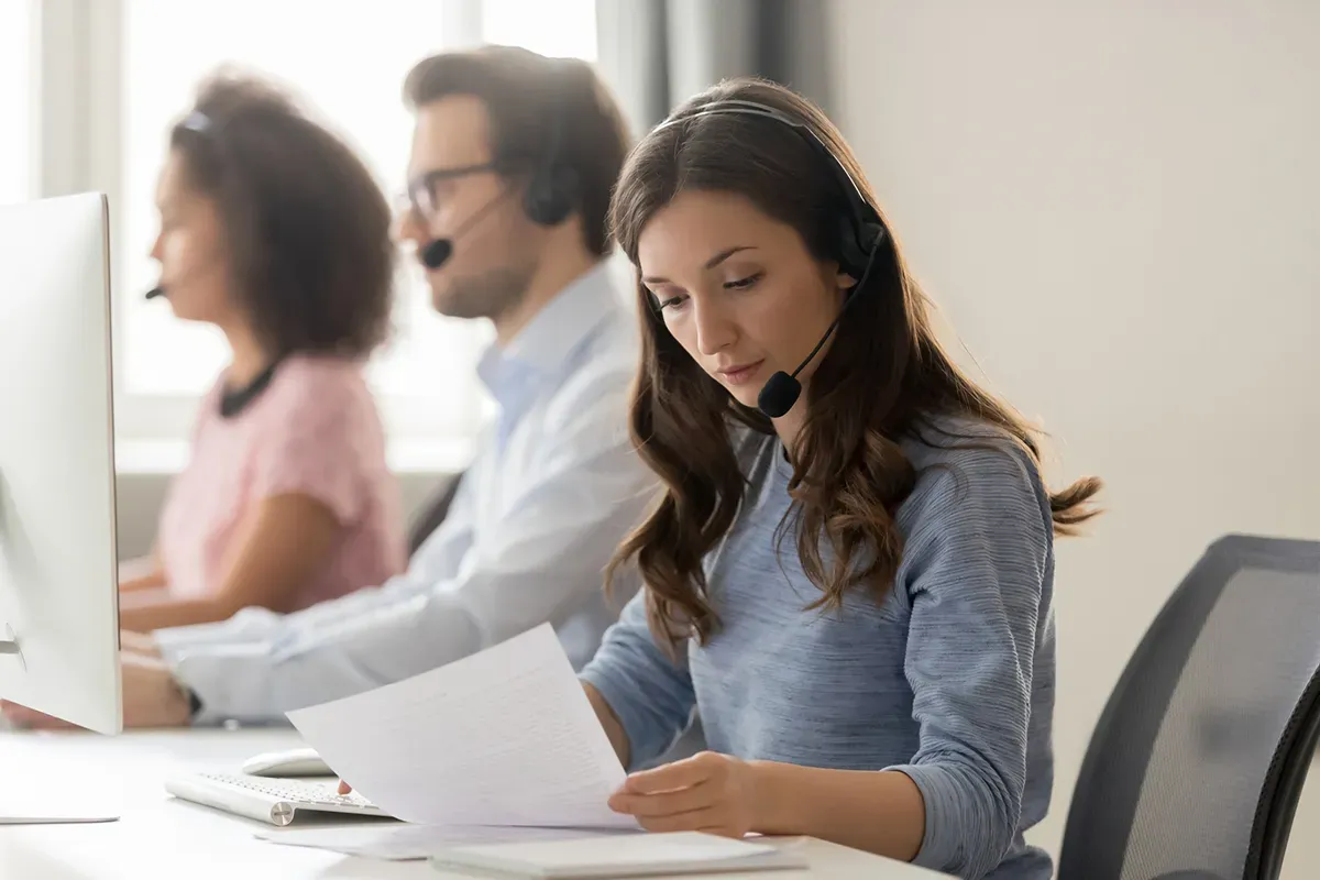 Three call center employees wearing headsets work at computers, with one focused on a document.