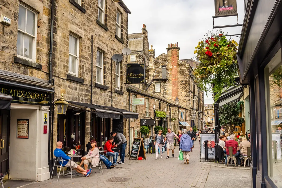 A quaint cobblestone street in Harrogate, lined with historic stone buildings, outdoor cafes, and people strolling or dining. Flower baskets adorn the scene.
