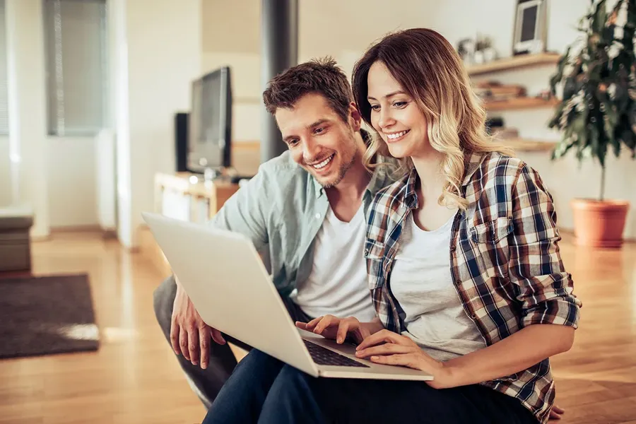 2 young homeowners sitting on their wooden floor of an open plan lounge watching a video on their laptop on how transferring equity works