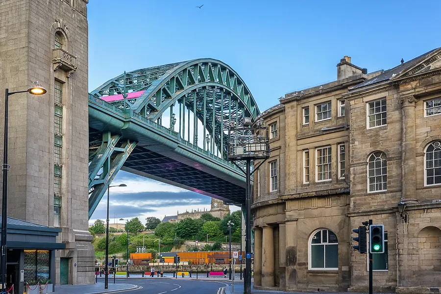 The iron Tyne bridge spans over a street in Newcastle, with the city skyline in the background. The structure features arches and connects buildings on either side, surrounded by old yellowstone buildings and a metropolitan landscape.