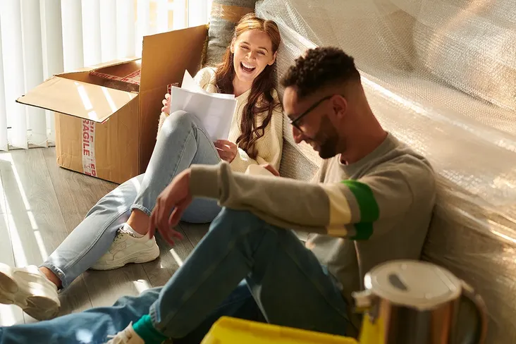 young couple sitting on the floor of their flat surrounded by boxes and a bubble wrapped sofa. Looking at their final moving paperwork and smiling at each other, excited to be moving to their new home