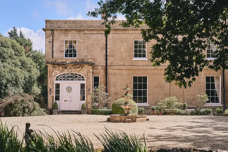 beautiful manor house front with large rectangular windows and white front door. There is an ornate fountain in the courtyard and sun is shining