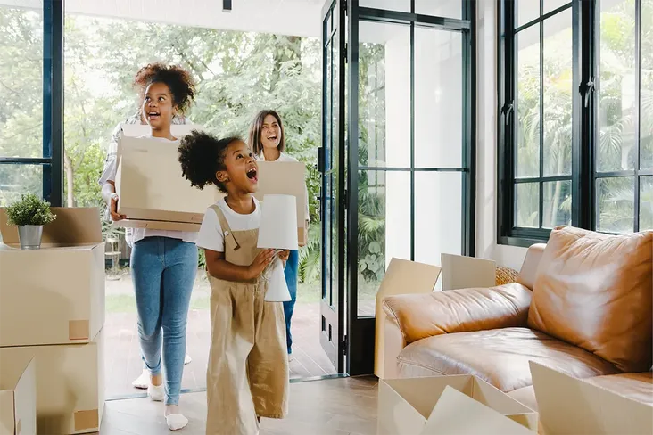 mum entering a new house with her 2 daughters all carrying boxes, ready to  move into their new home. The house is bright and with floor to ceiling windows and the children look excited