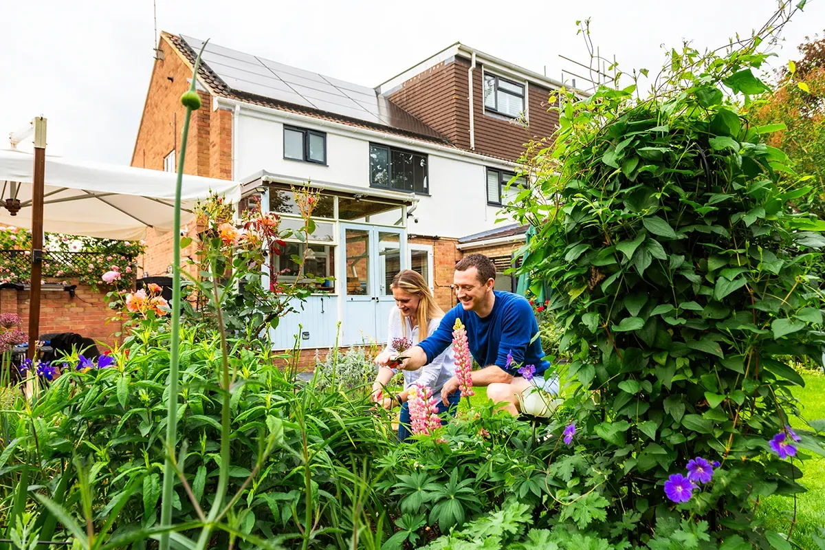 young couple kneeling in their back garden and digging to add in more flowers. The garden is established with lots of plants and the house is in the background