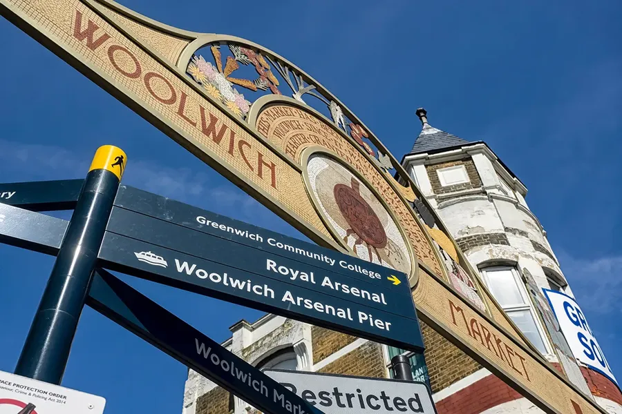 historical sign for woolwich market with a sign post next to it showing directions to woolwich arsenal and the pier