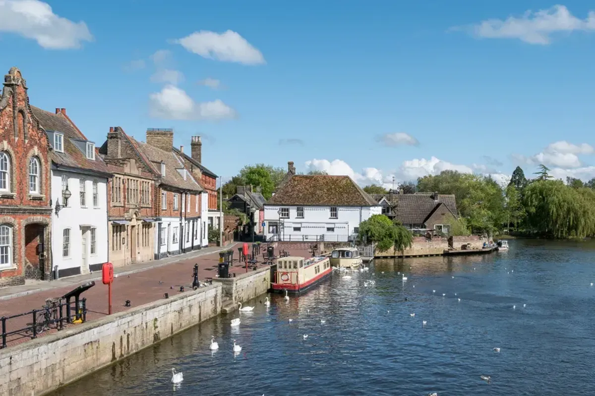A scenic riverside scene with traditional brick buildings, a docked boat, and swans on a sunny day under a blue sky with fluffy clouds.