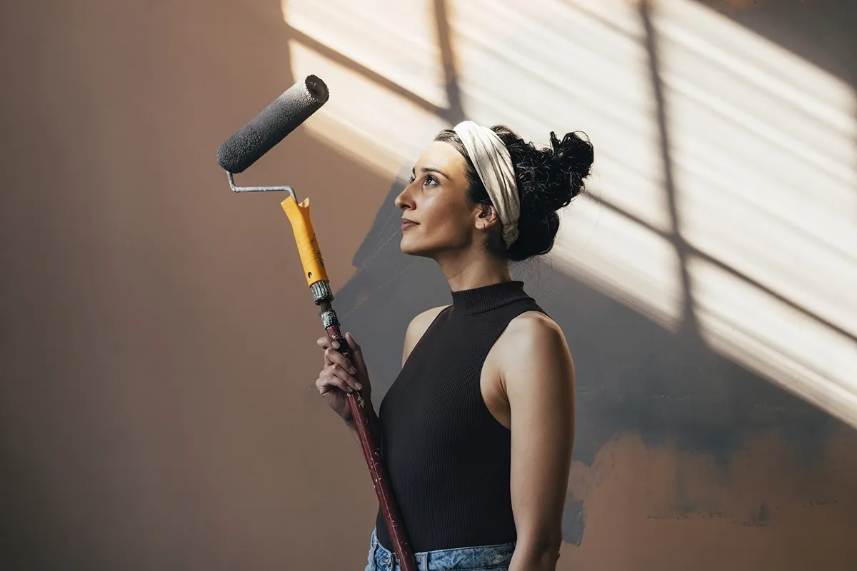 Woman with a headband holding a paint roller, looking at a sunlit wall with painted patches, wearing a sleeveless top and jeans.