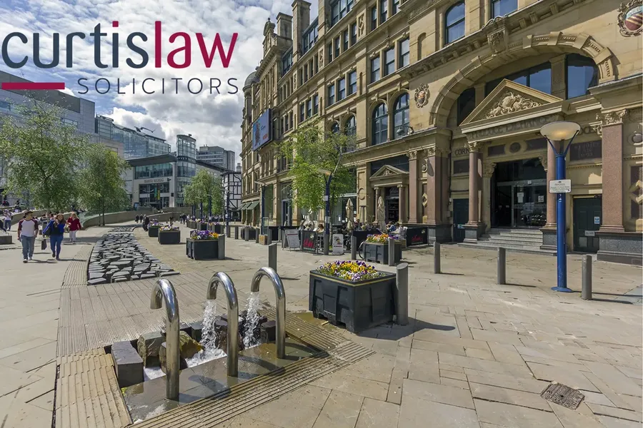 Manchester city street scene with historic buildings, modern office, and small fountain. People walking and flower planters under a partly cloudy sky.
