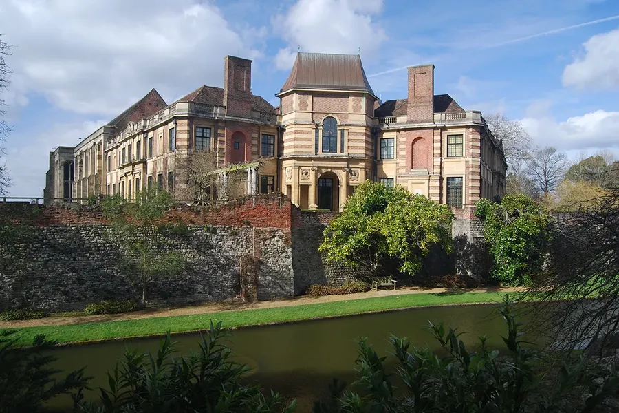 Historic Eltham Palace, a brick mansion with a central tower, surrounded by a stone wall and moat, set against a partly cloudy sky.
