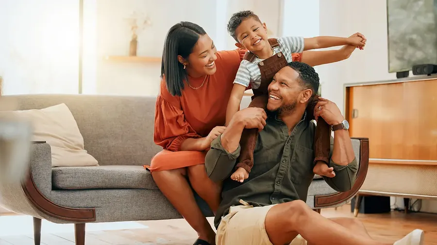 A cheerful family of three in a living room, with the child playfully sitting on the father's shoulders and the mother smiling at them.