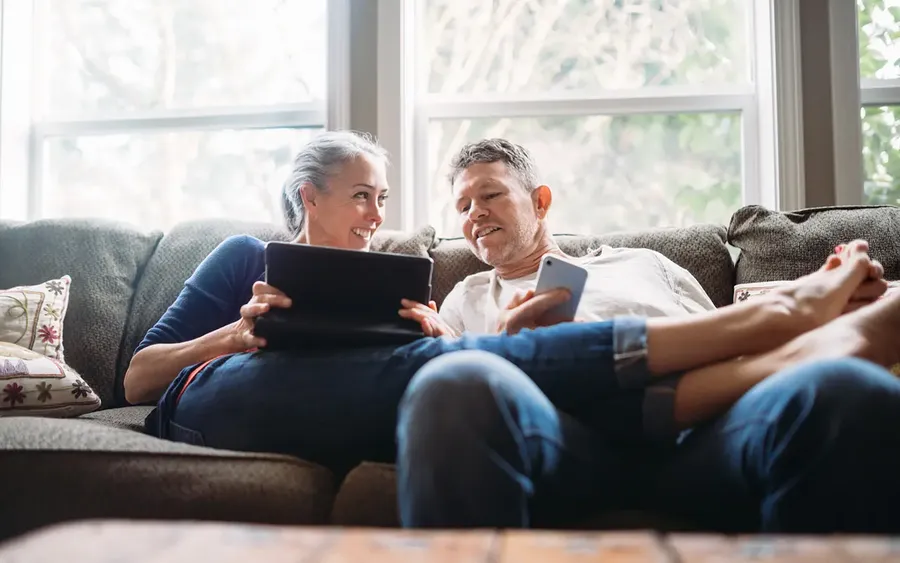 An older couple relaxes on a sofa, smiling at each other. One holds a tablet, and the other holds a smartphone. Sunlight filters through the window.