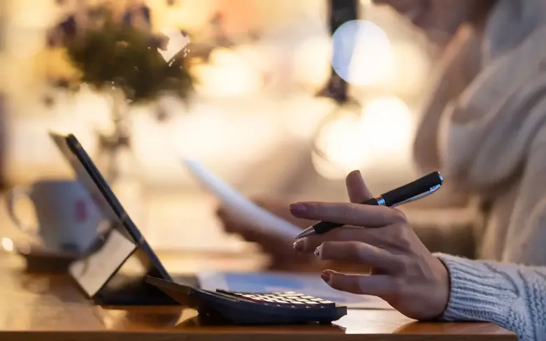 Person holding a pen, using a laptop and calculator at a wooden table, with a mug and papers, in a softly lit setting.