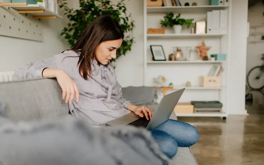 Young woman sitting on a couch using a laptop in a cozy living room with plants and shelves in the background.