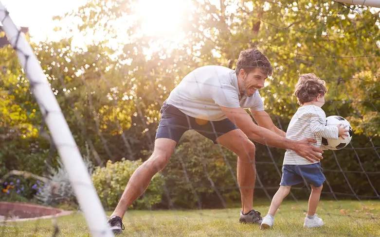 Man and child playing soccer in a sunny garden. The man is playfully reaching for the ball as the child runs with it.
