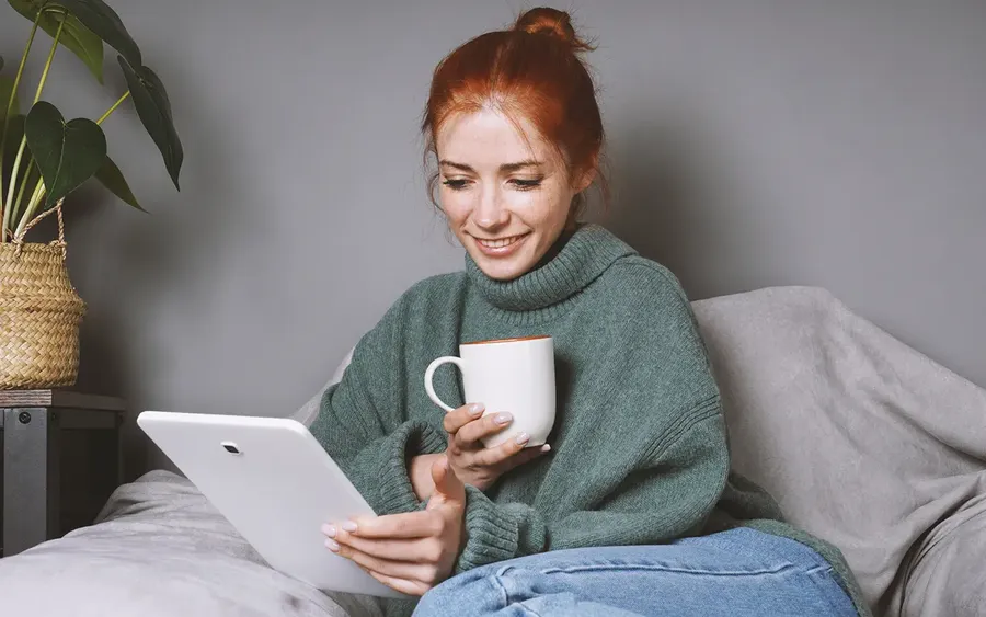 A woman with red hair sits on a sofa, smiling, looking at a tablet. She wears a green sweater and holds a white mug. A potted plant is on a table beside her, and the background is a neutral grey wall.