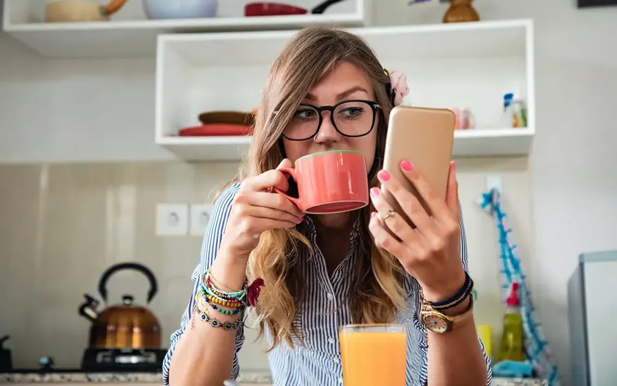 Woman with glasses sips from a red mug while looking at her phone in a kitchen. A glass of orange juice is on the counter.