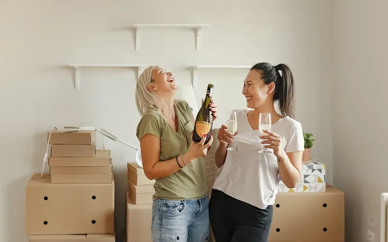 young female couple celebrating, laughing and drinking champagne in their new home