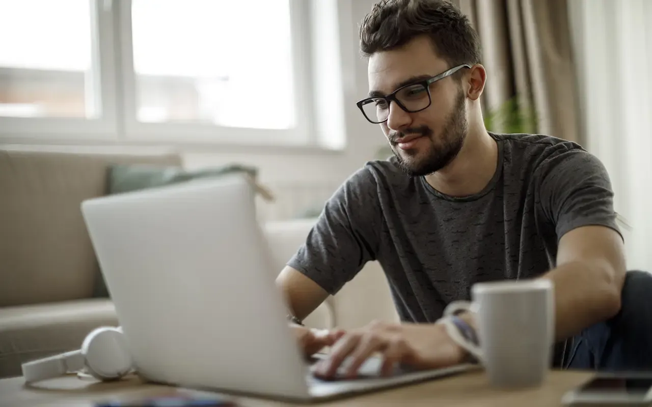 Young man with glasses typing on a laptop at a table, with a coffee mug and headphones nearby, in a well-lit room.