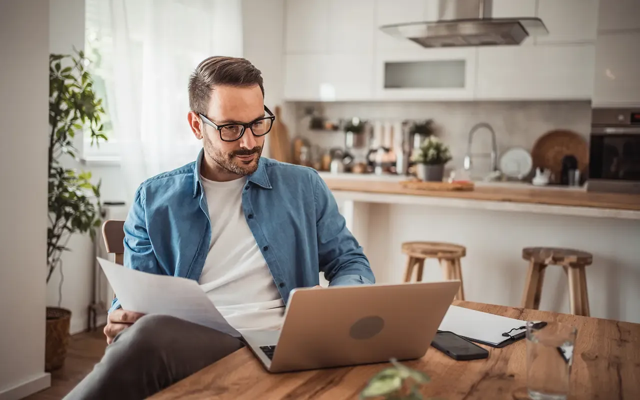 Man with glasses working on a laptop at a wooden table, holding papers, in a modern kitchen setting with bright natural light.