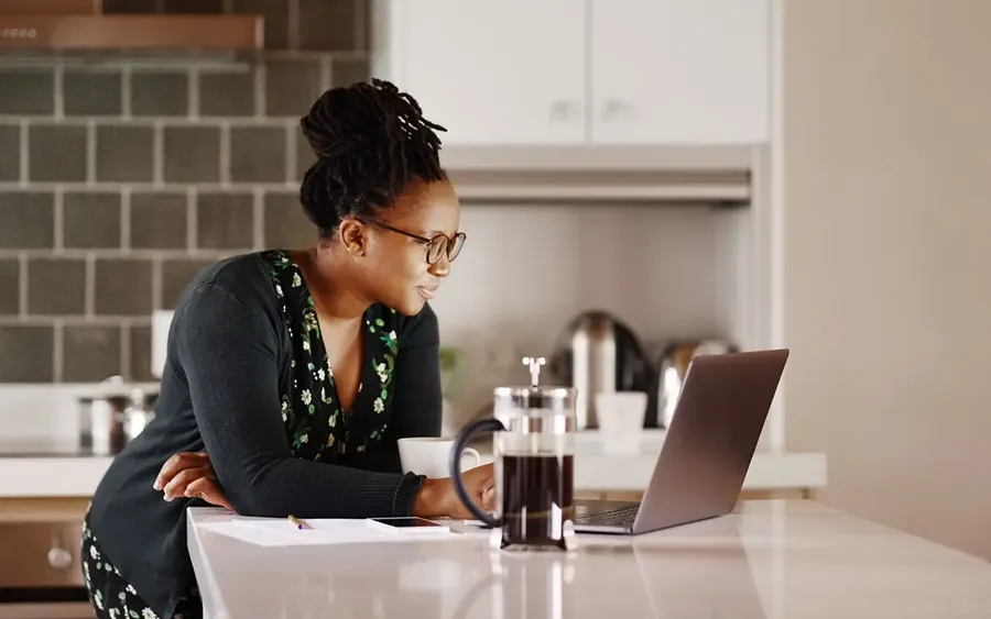 Woman with glasses and braided hair, wearing a dark sweater, working on a laptop at a kitchen counter with a French press and papers nearby.