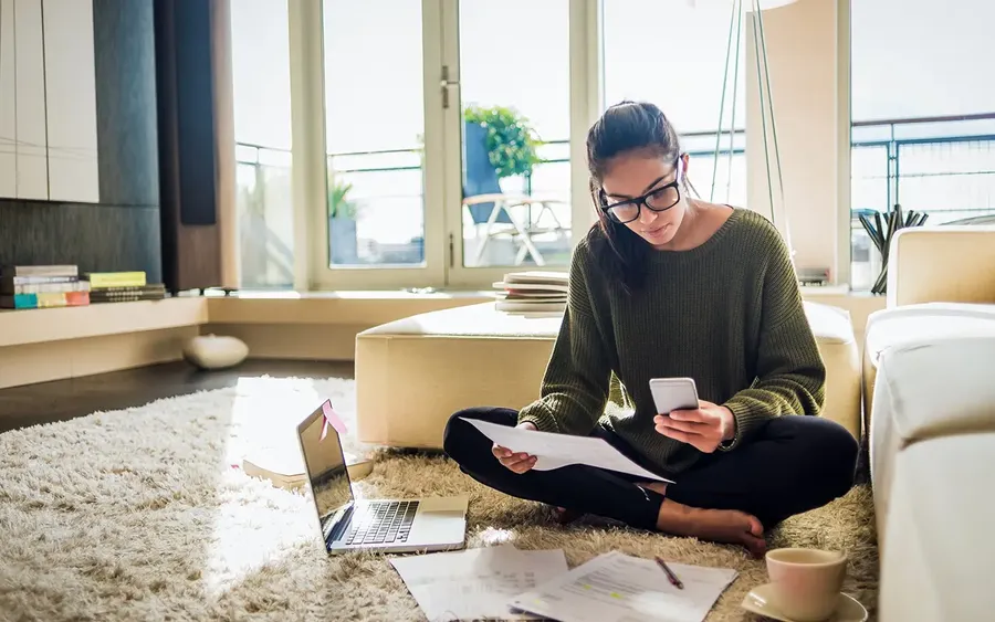 Woman sitting on a rug, using a smartphone and reading documents, with a laptop nearby in a bright, modern living room.