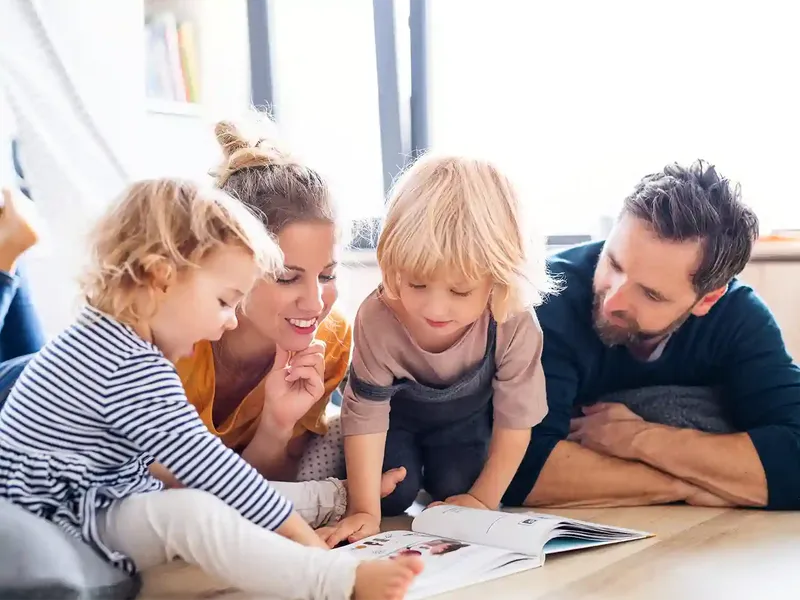 A family of four, including two young children, is lying on the floor, engaged in reading a book together in a bright room.
