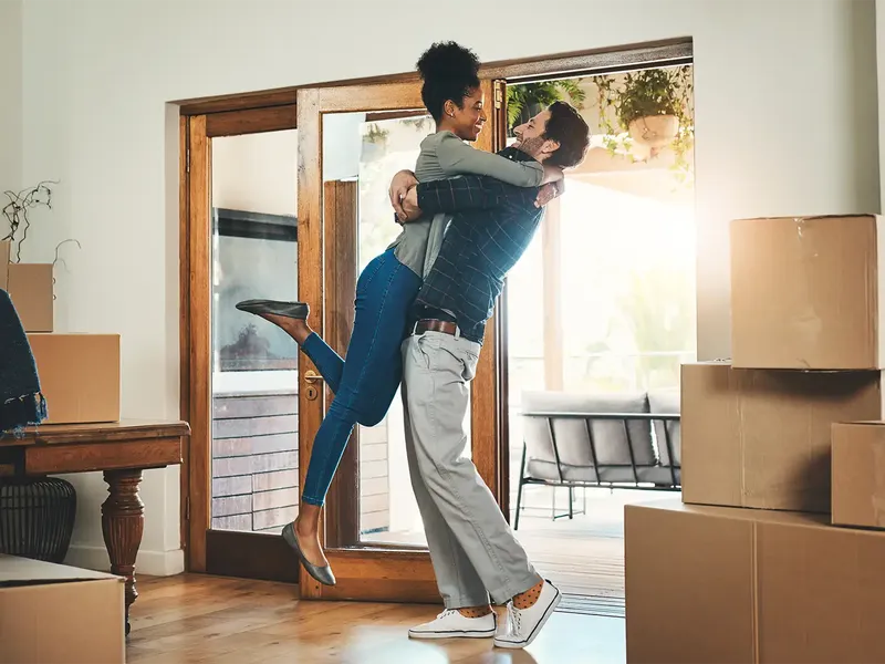 A joyful couple embrace in a sunlit room surrounded by cardboard boxes. The woman is lifted off her feet by the man. They are moving into a new home, with sunlight streaming in through large windows.