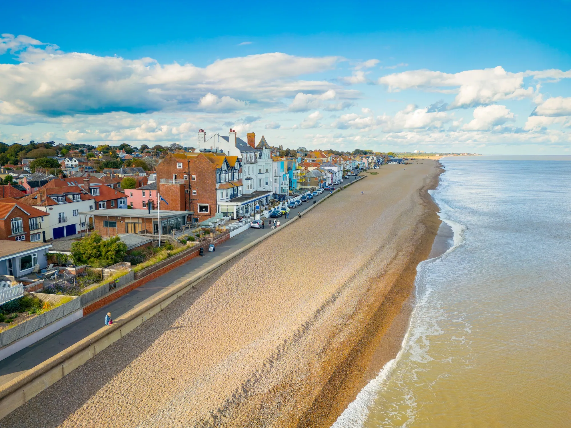 Seaside town with various houses lining a long beach, calm sea waves, and a pathway for walking under a clear blue sky with white clouds.